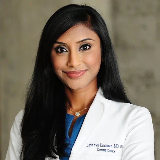 A person with long dark hair wearing a white lab coat over a blue top, standing indoors.