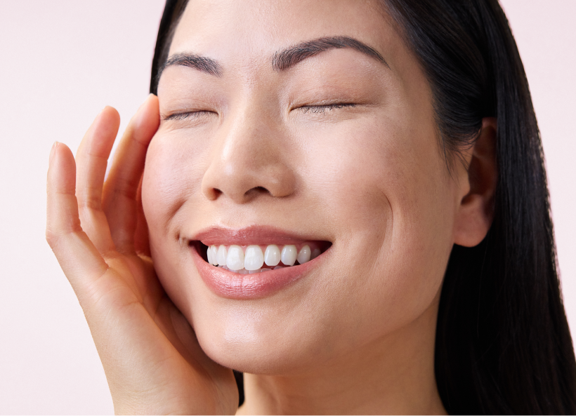 A person with long dark hair smiles with eyes closed, showing teeth, and lightly touches their face.